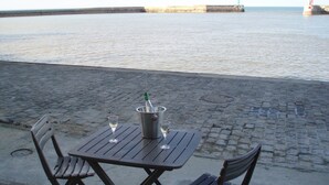Ein Tisch mit zwei Stühlen und einer Champagnerflasche auf einem bepflasterten Bereich in der Nähe von Wasser.