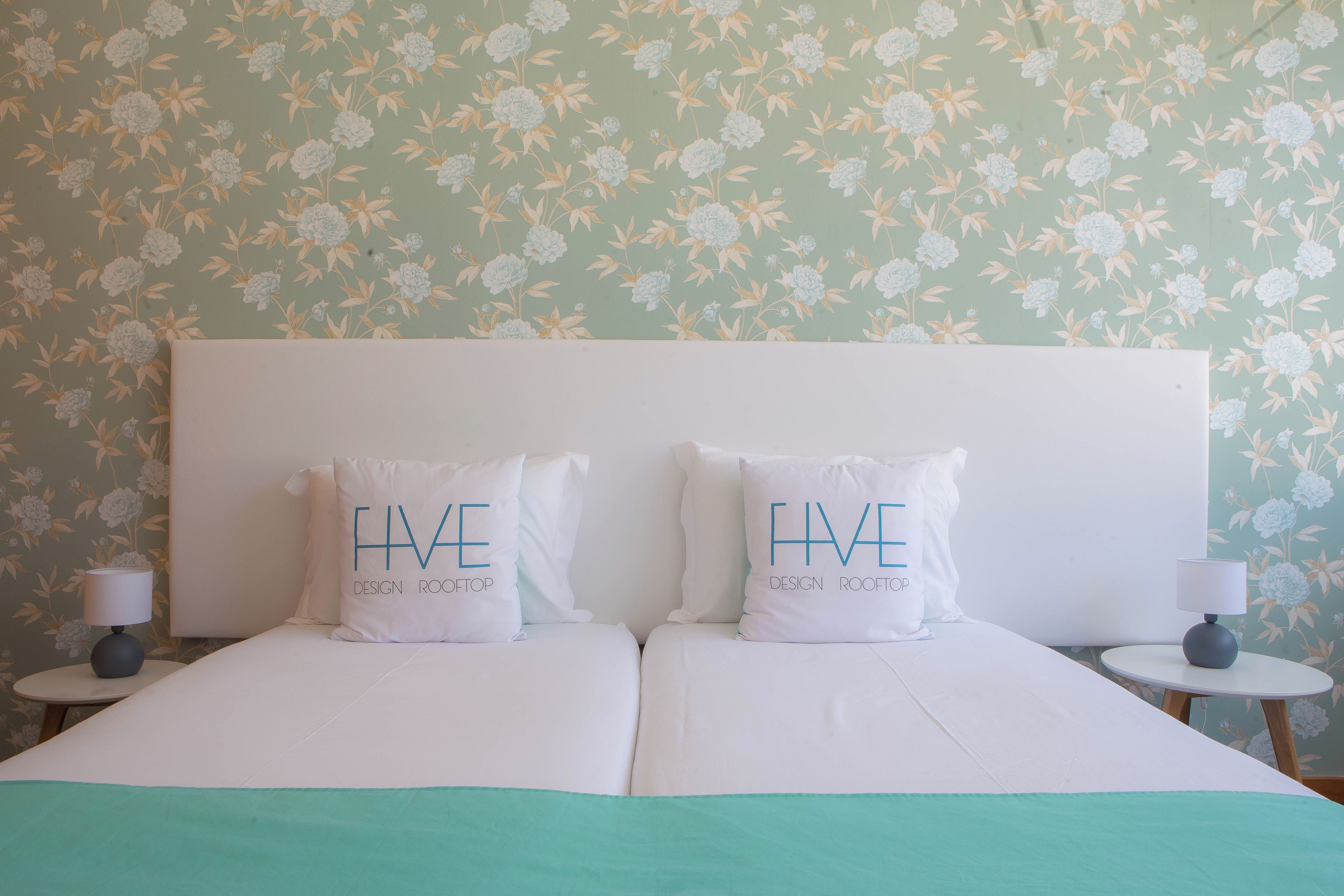 A double bed with two pillows, a nightstand, a lamp, and a floral wallpaper.