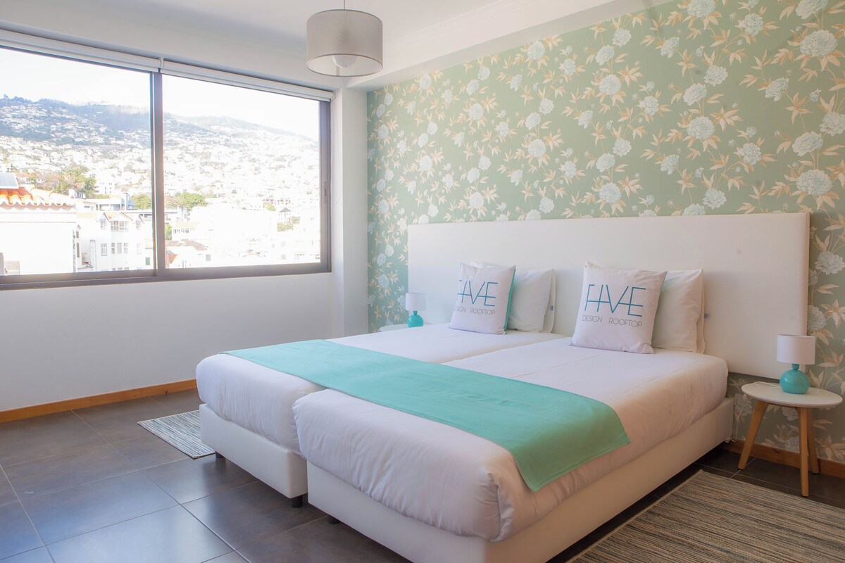 Un lit double avec une couvre-lit de couleur turquoise et deux oreillers, une petite table de chevet avec une lampe, et une vue sur le paysage urbain par une grande fenêtre.
