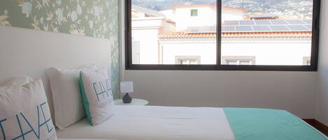 Une chambre avec deux lits, une fenêtre donnant sur des immeubles et des montagnes, et un papier peint à motifs floraux.