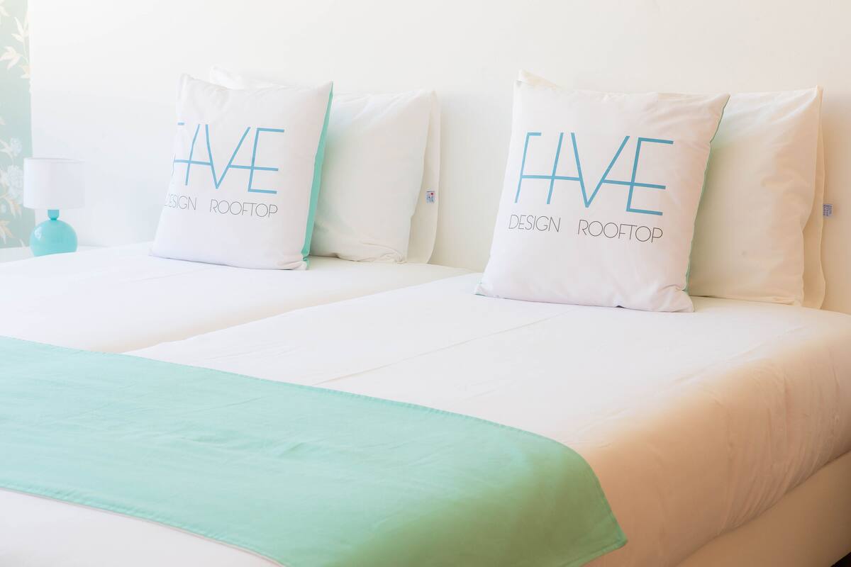 Habitación de hotel con dos camas, cada una con un cojín que dice "HAVE DESIGN ROOFTOP".