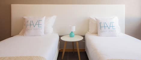A hotel room with two single beds, each with a pillow that reads "HAVE DESIGN ROOFTOP". There is a small round table with a lamp between the beds.