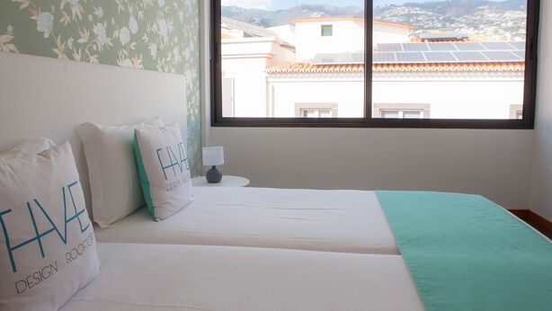 A bedroom with two beds, a window with a view of buildings and mountains, and floral wallpaper. - Five Design Rooftop by Storytellers (Funchal)