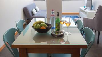 Una mesa de comedor con un frutero, botellas de vino y copas, rodeada de sillas color turquesa en una habitación moderna con un sofá y un escritorio al fondo.