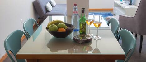Une table à manger avec un panier de fruits, des bouteilles de vin et des verres, entourée de chaises turquoise dans une pièce moderne. On aperçoit un canapé et un bureau à l’arrière-plan.