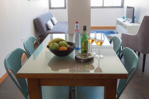 Spisebord med skål med frugt, vinflasker og glas, omgivet af turkise stole i et moderne rum med en sofa og et skrivebord i baggrunden.