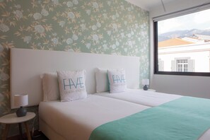 A double bed with two pillows, a bedside table with a lamp, and a window with a view of buildings and mountains. - Five Design Rooftop by Storytellers (Funchal)