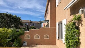 A courtyard with a paved walkway, a building with a beige facade, and some greenery. - SEASONAL RENTAL - LOWER VILLA (La Londe-Les-Maures)
