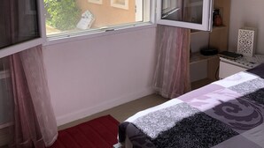 A bedroom with a large window, a bed, a bedside table, a bookshelf, and a red carpet. - SEASONAL RENTAL - LOWER VILLA (La Londe-Les-Maures)