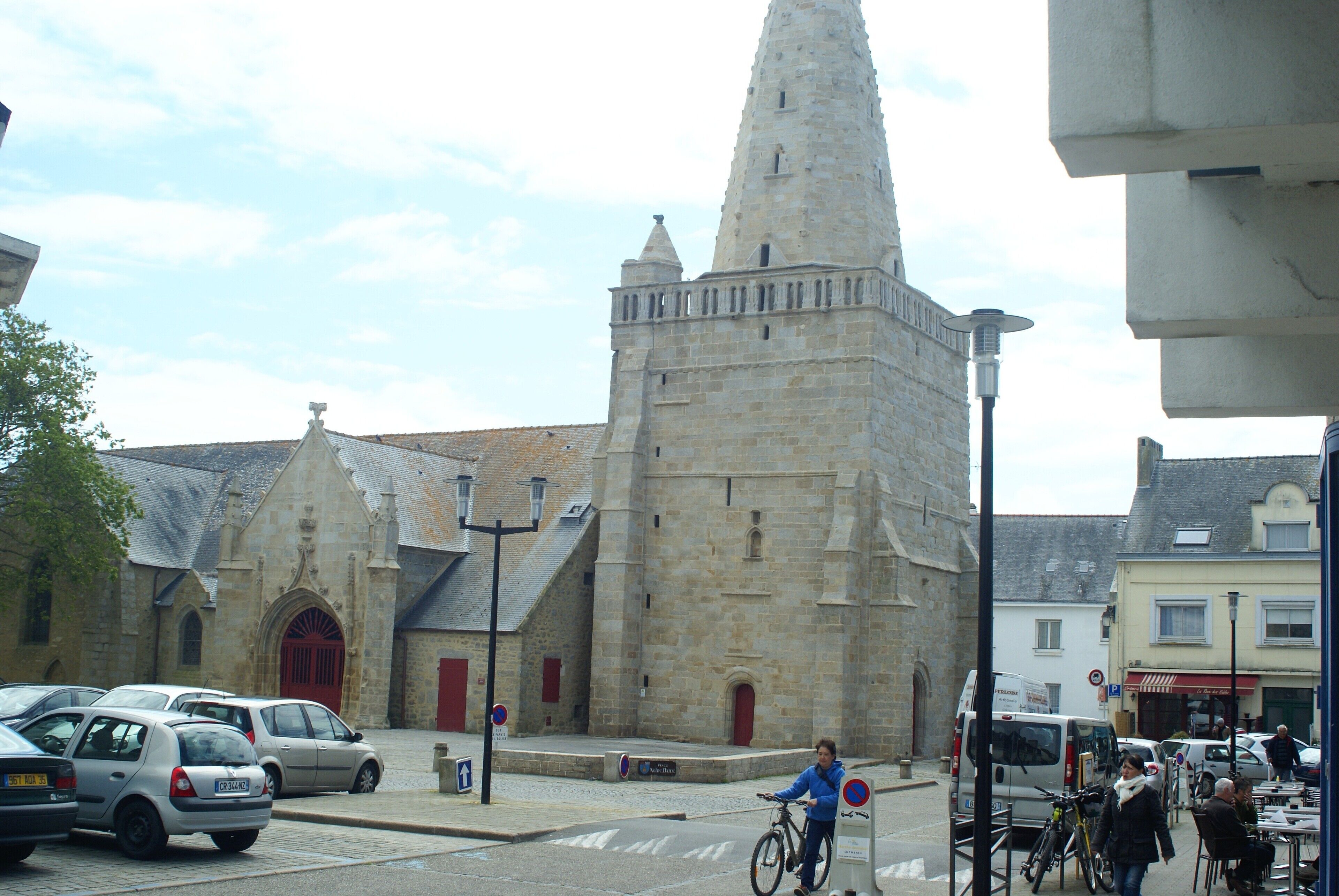 A historic stone building with a prominent tower, a paved street, parked cars, and a person riding a bicycle.