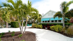 A house with a white exterior and blue awnings, surrounded by lush greenery and a gravel driveway. - Welcome to Beach Basket – Your Seaside Retreat!" (Anna Maria)