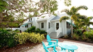 A two-story house with a porch, a staircase, and a garden with various plants and a blue Adirondack chair. - Welcome to Beach Basket – Your Seaside Retreat!" (Anna Maria)