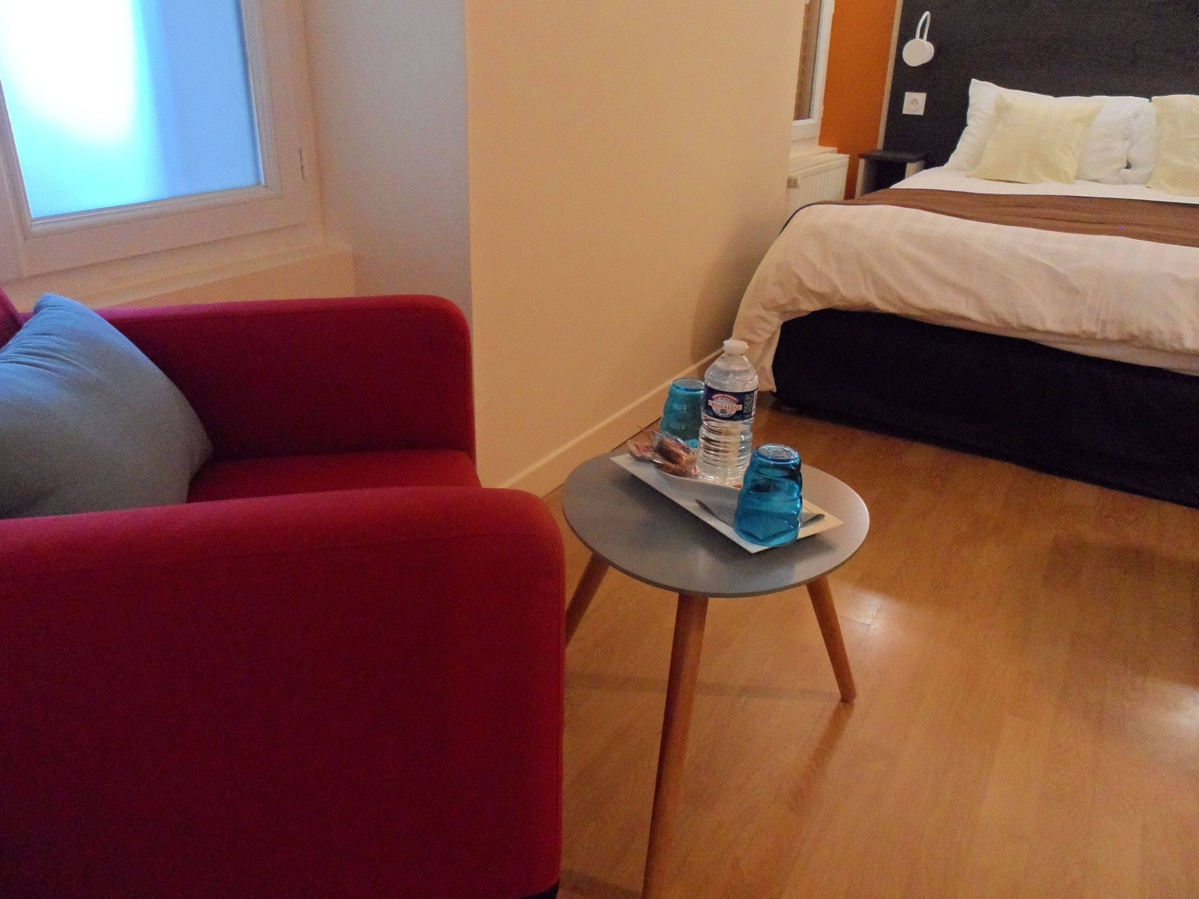A hotel room with a red armchair, a small table with a tray of snacks, a bottle of water, and a glass of water.