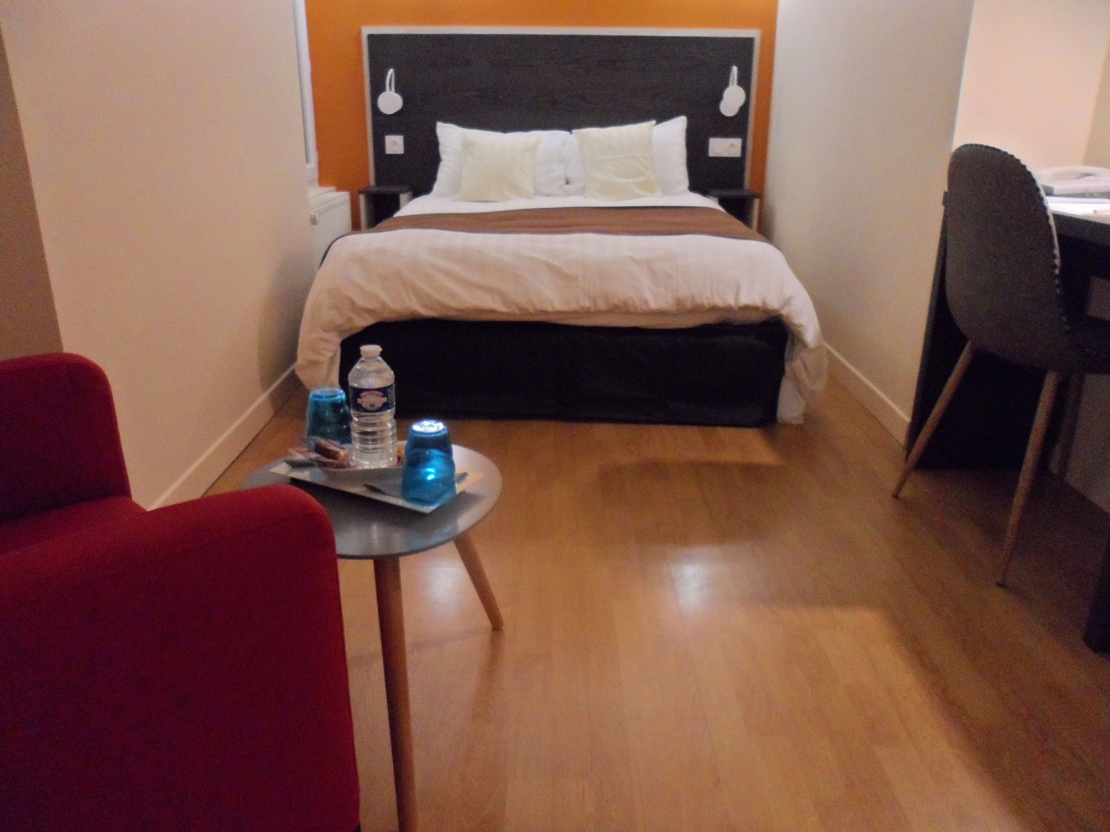 Een compacte hotelkamer met een bed, een klein tafeltje met een dienblad water en snacks, een rode stoel en een bureau met stoel.