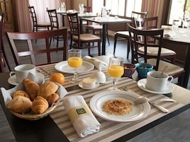 Mesa de café da manhã com croissants, suco de laranja, café e uma tigela de frutas.