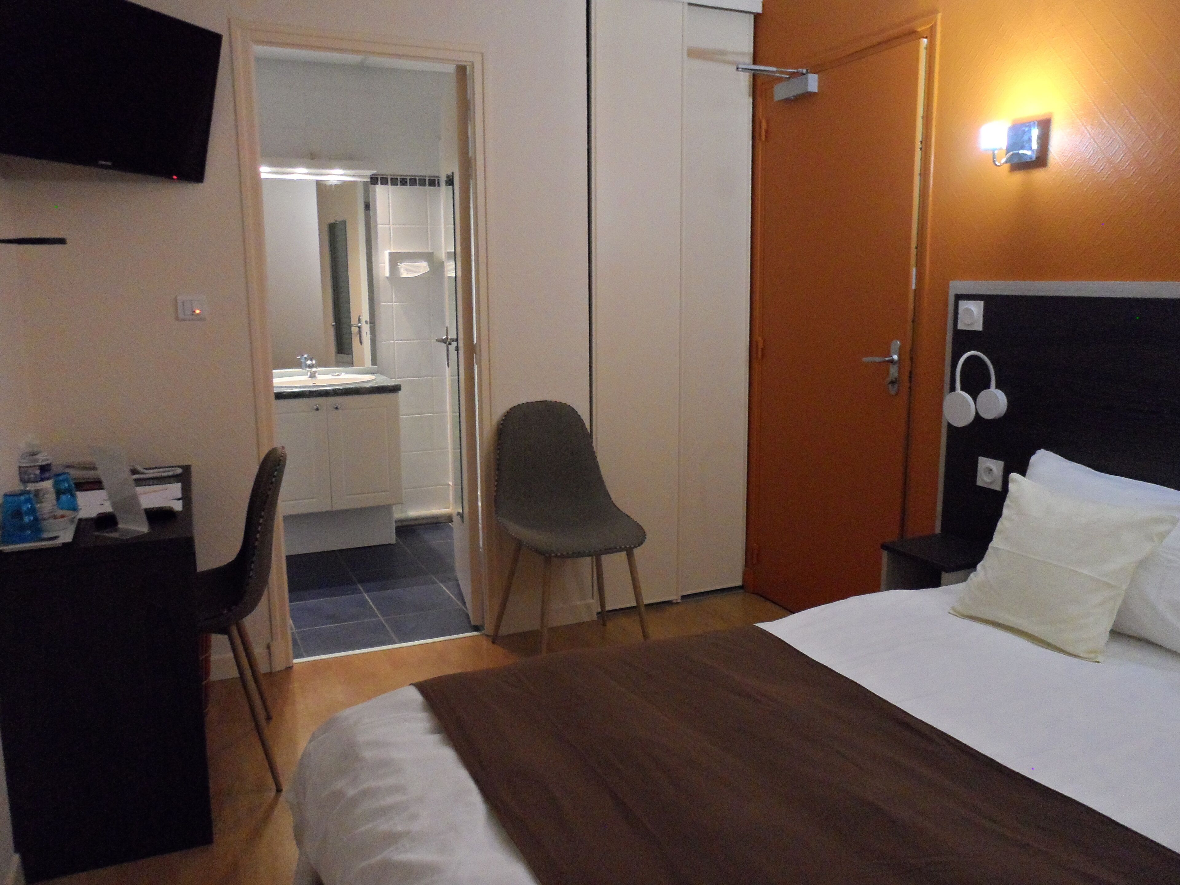 A hotel room with a bed, a desk with a chair, a bathroom visible through an open door, and a wall-mounted TV.