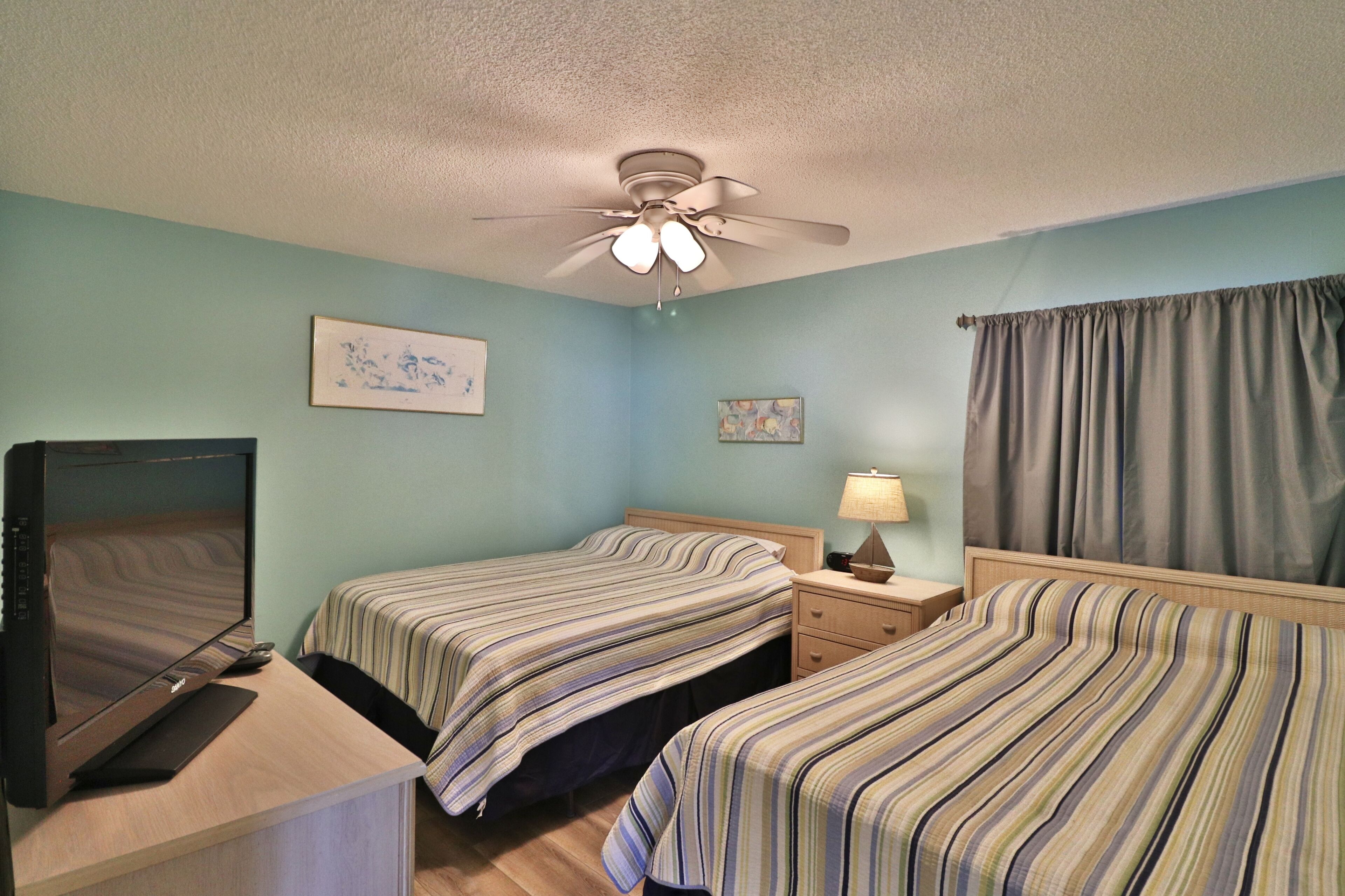 A hotel room with two beds, a desk with a computer, a ceiling fan, and a window with curtains.