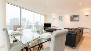 Uma sala de estar moderna com uma mesa de jantar de vidro, cadeiras estofadas cinzentas, um sofá de couro preto e uma grande janela com vista para a cidade.