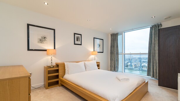 Quarto de hotel moderno com uma cama grande, duas mesas de cabeceira com luminárias, vista da cidade através de janelas amplas e obras de arte emolduradas nas paredes.