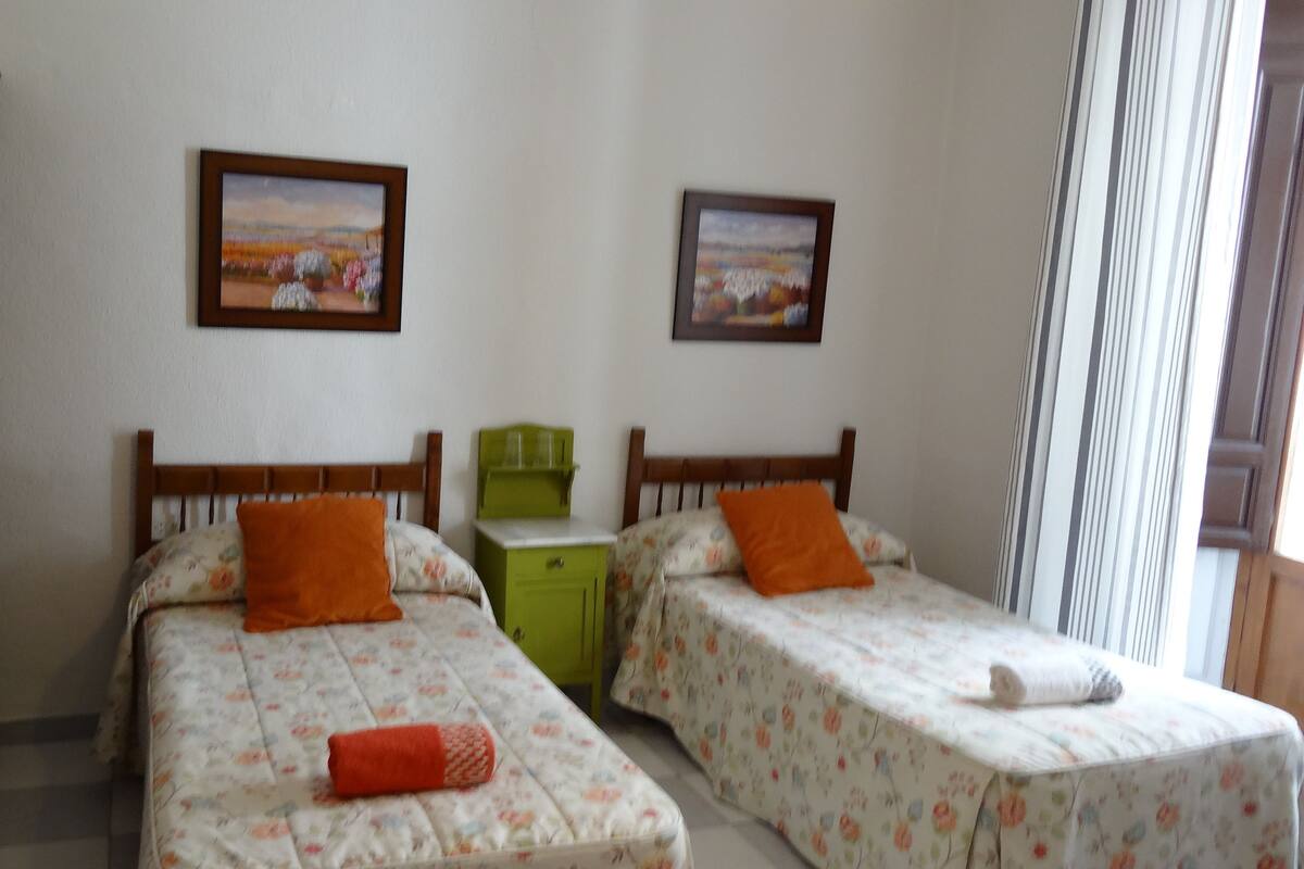A room with two single beds, each with a floral bedspread and an orange pillow. There is a green nightstand with a glass set on top. Two framed pictures are hung on the wall above the beds.
