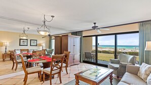 A spacious living area with a dining table, chairs, and a sofa. There is a view of the beach through large windows.