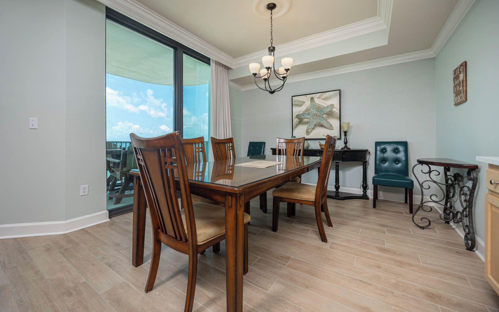 A dining area with a glass table and wooden chairs, a chandelier, and a view through large windows.