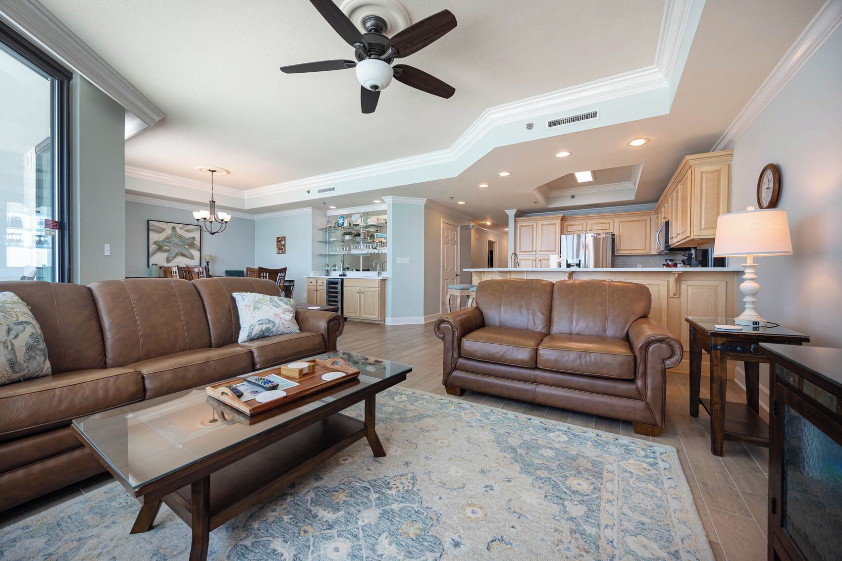 A spacious living room with a brown leather sofa, a matching armchair, and a coffee table with books and a tray. A ceiling fan and recessed lighting are visible.