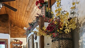 A rustic interior with wooden floors, a ceiling fan, a wooden dresser with decorative items, a wall-mounted basket with flowers, and a shelf with ceramic dishes. - RIVERFRONT ADIRONDACK LOG CABIN (Warrensburg)