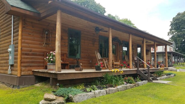 A log cabin with a porch, wooden chairs, and flower pots. - RIVERFRONT ADIRONDACK LOG CABIN (Warrensburg)
