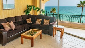 A living room with a dark brown sectional sofa, two wooden coffee tables, and a large window overlooking the sea. - Sonoran Spa W403 - 2 BR Next to the Pool, High Speed Wi-Fi - Beachfront Upper Floor Luxury Condo (Puerto Peñasco)