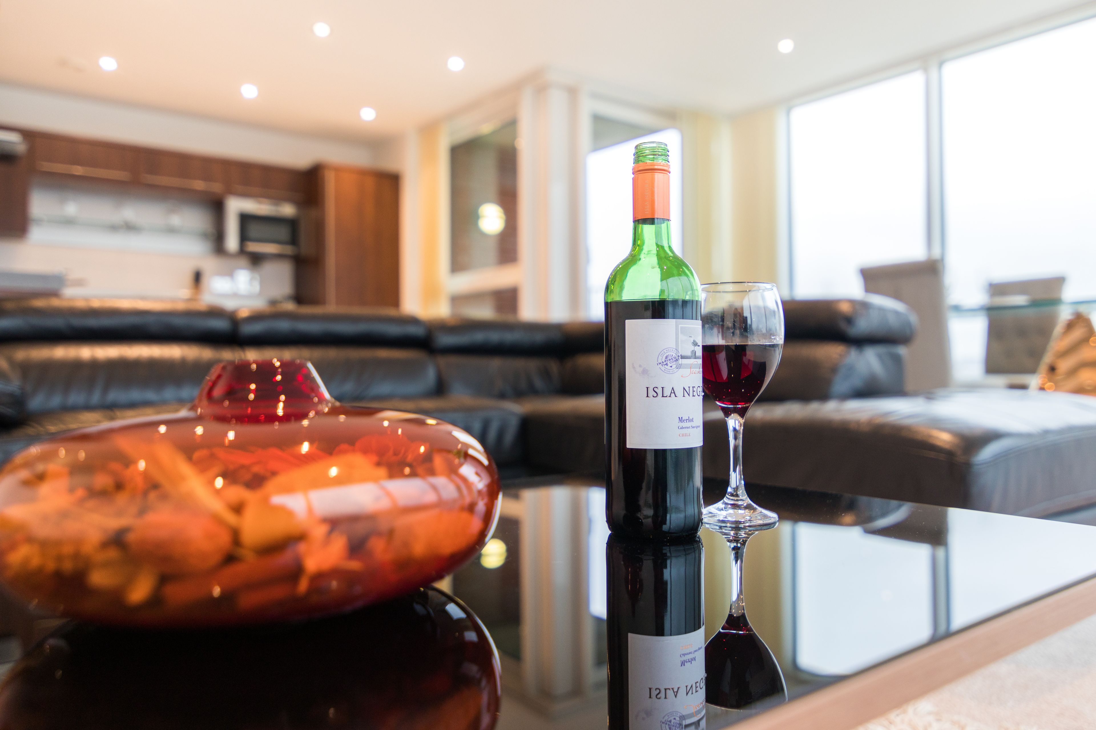 Een fles wijn en een glas op een tafel in een moderne woonkamer met een leren bank en een keuken op de achtergrond.