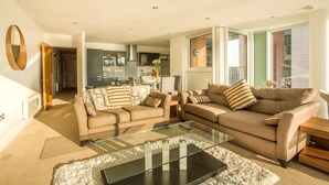 A modern living room with two sofas, a glass coffee table, and a kitchen in the background.