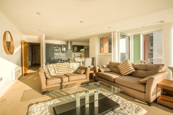 A modern living room with two sofas, a glass coffee table, and a kitchen in the background.