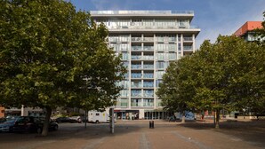 A modern building with multiple floors and balconies, surrounded by trees and a paved area.