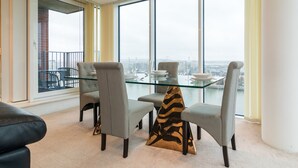 A dining area with a glass table and four grey chairs, a zebra-patterned cushion, and a city view through large windows.