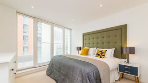 A bedroom with a large bed, a nightstand, and a window with blinds. - Grand Vista by Austin David Apartments (London)