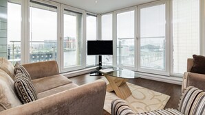 A modern living room with a glass coffee table, beige sofas, and large windows offering a city view. - Grand Vista by Austin David Apartments (London)