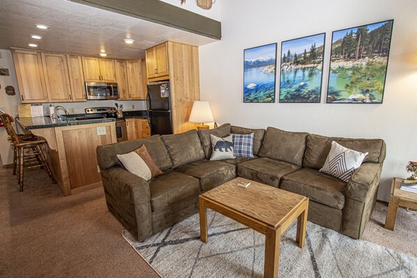 A living room with a brown sectional sofa, a wooden coffee table, and a kitchen area with wooden cabinets and appliances.