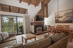 A spacious living room with a fireplace, wooden furniture, and a large window overlooking a deck.