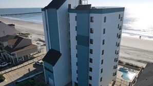 Ein mehrgeschossiges Hotel mit Pool im Erdgeschoss, nahe einem Strand mit einem Pier im Hintergrund.