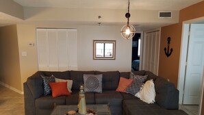Ein Wohnzimmer mit einem dunkelgrauen Ecksofa, einem Glastisch mit Dekorationen, einer Hängeleuchte und einer Tür mit Ankerverzierung.