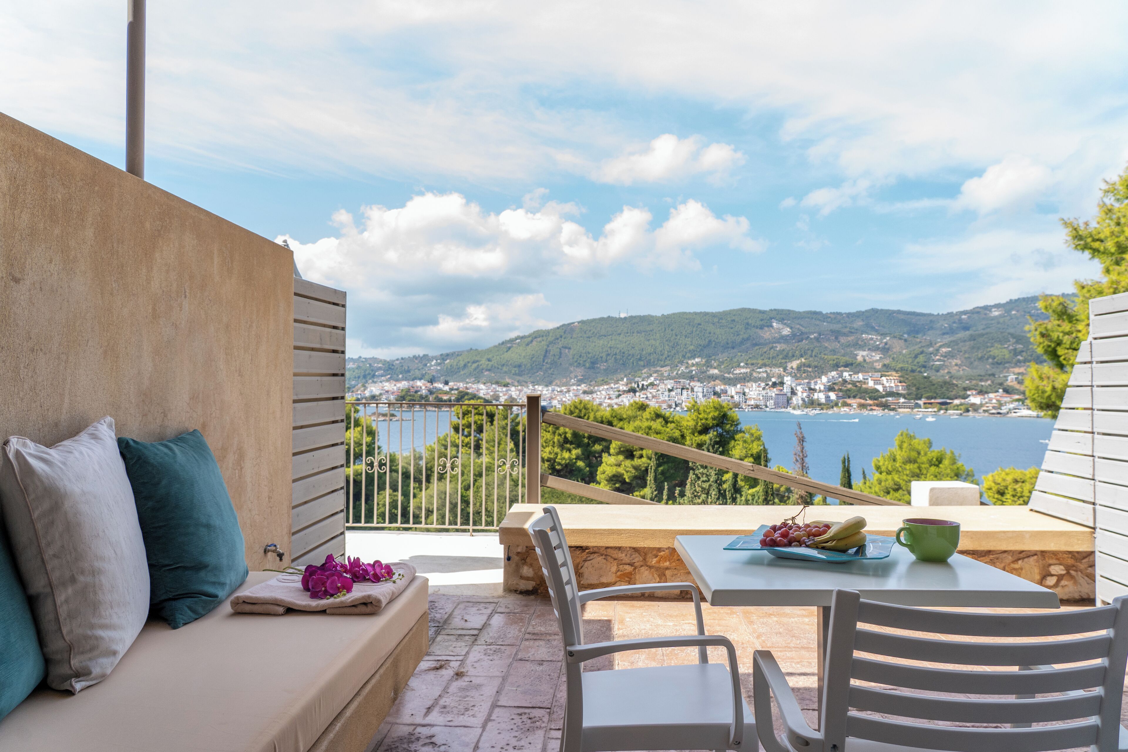 A balcony with a sofa, chairs, and a table with a view of a coastal town and hills.
