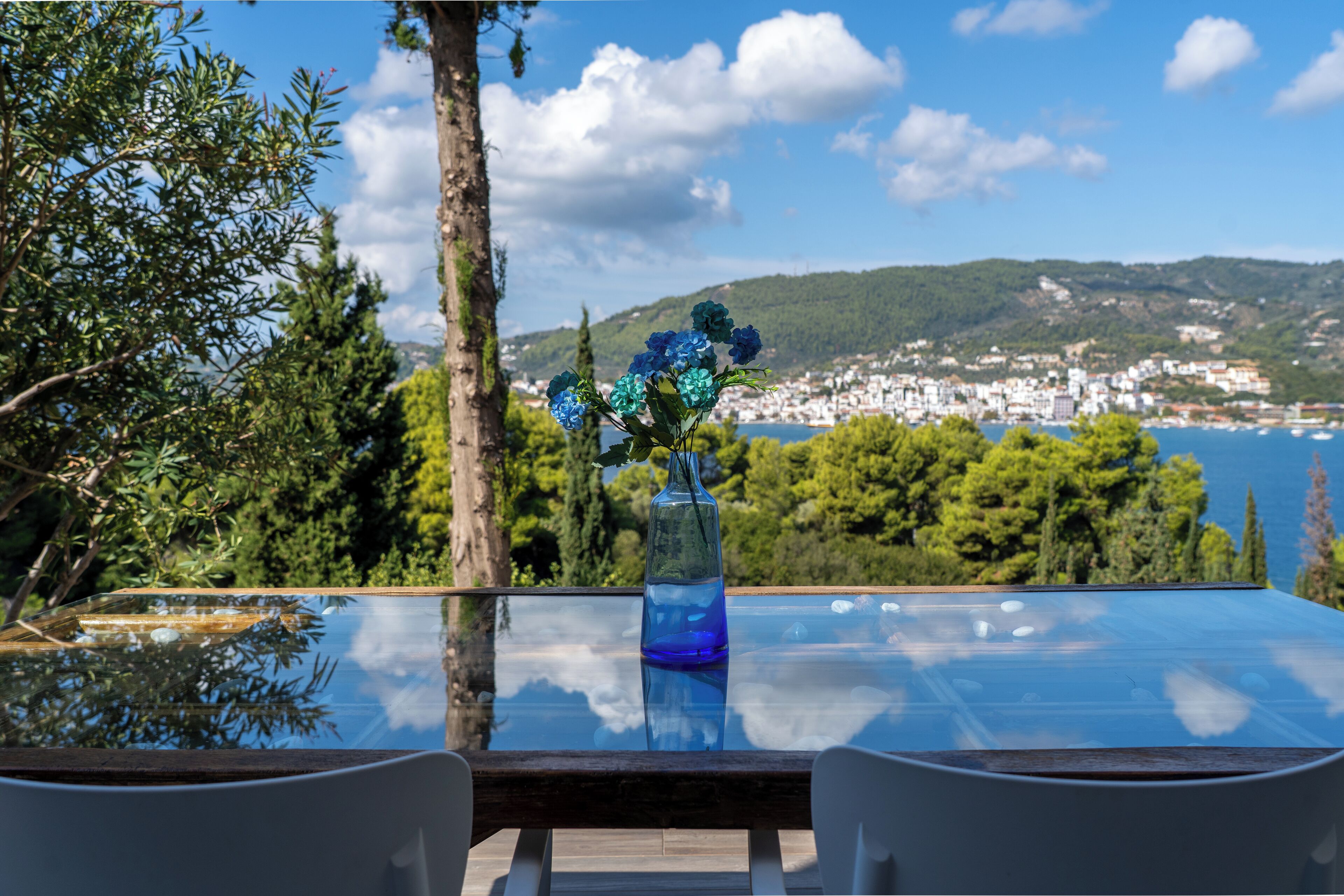 A glass table with a blue vase and white chairs, overlooking a coastal town and hills.