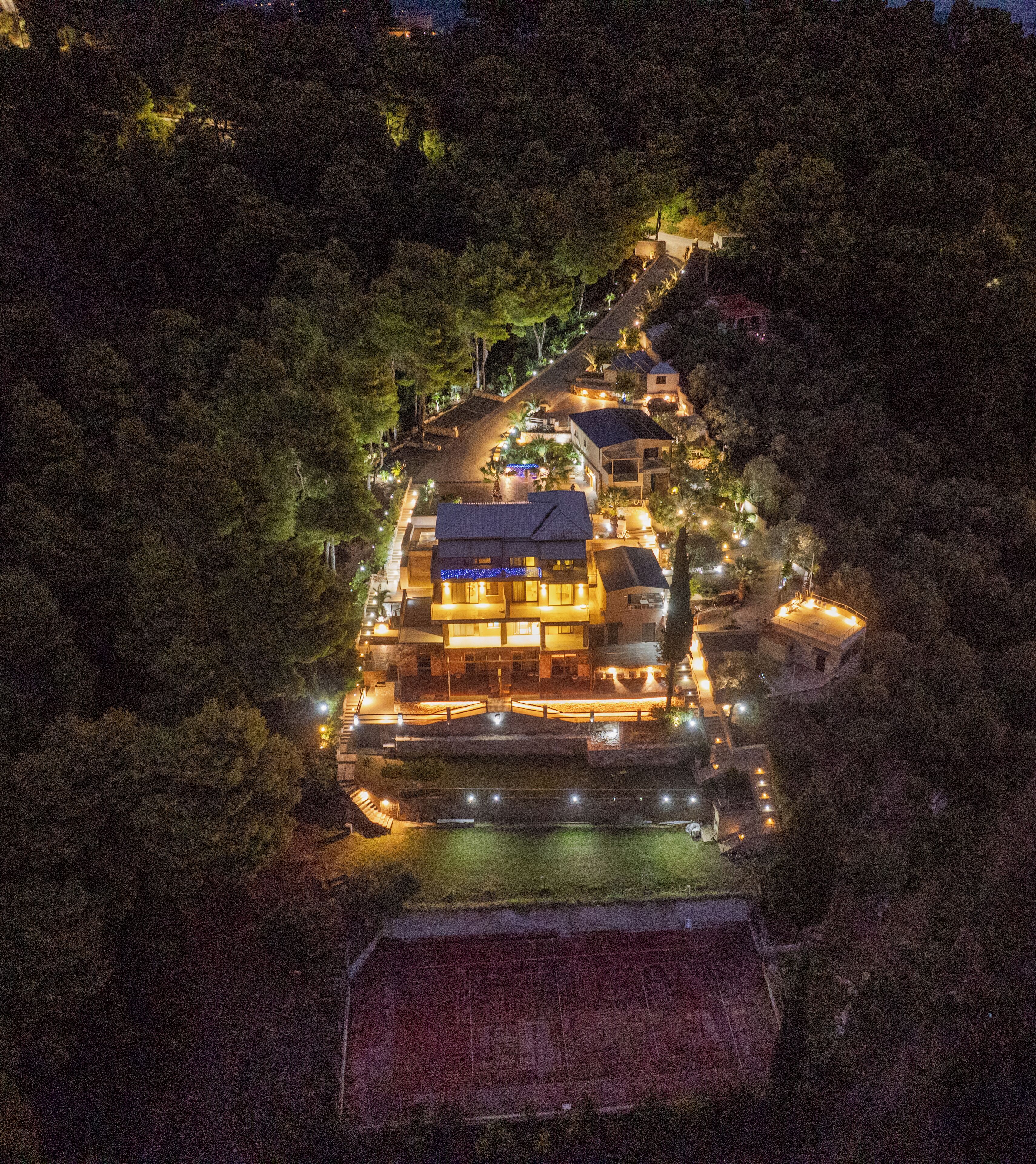 An aerial view of a hotel surrounded by dense forest, featuring a central building with multiple floors and a well-lit exterior.