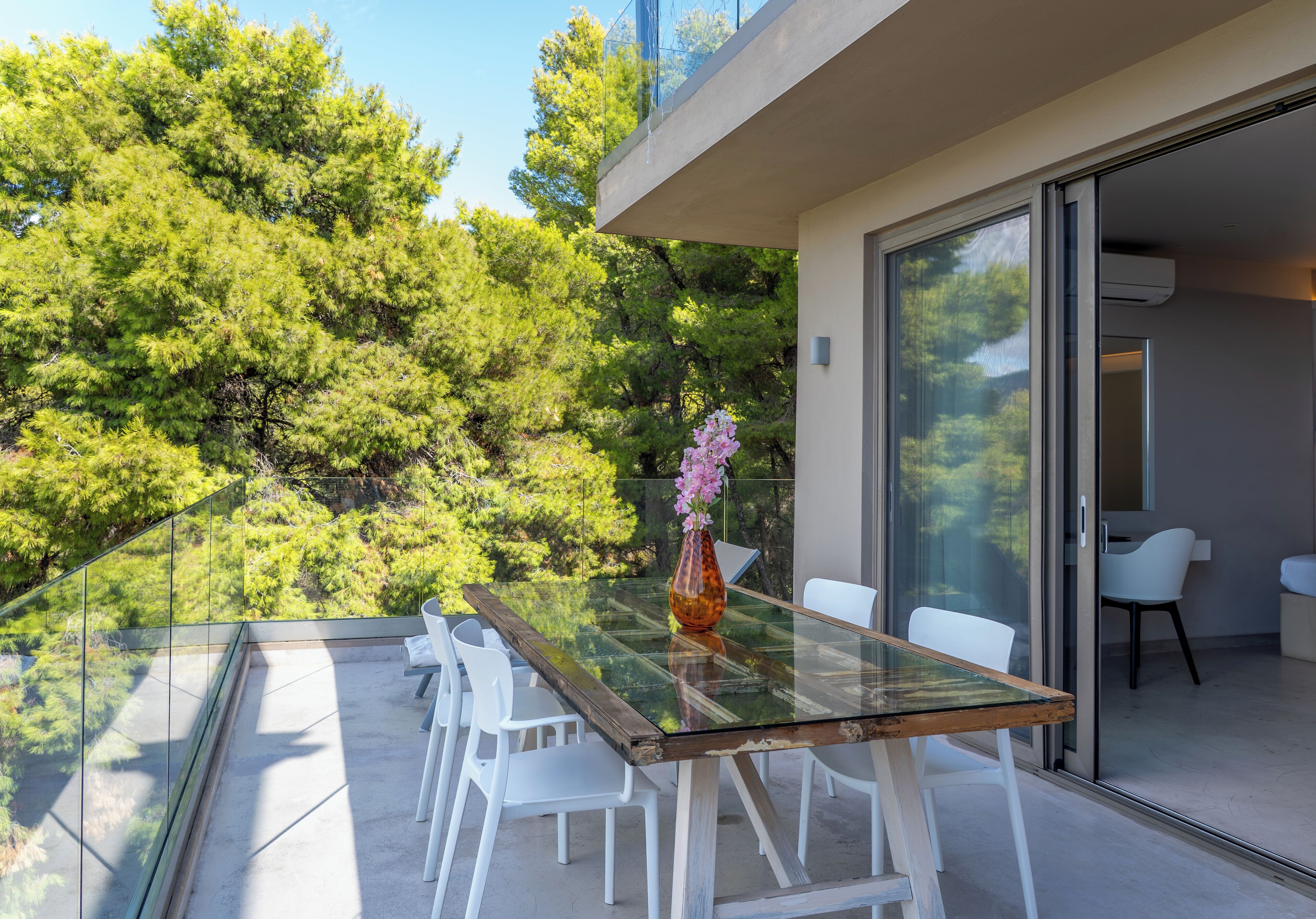 A modern outdoor dining area with a glass table and white chairs, overlooking a lush garden and a building with large glass doors.