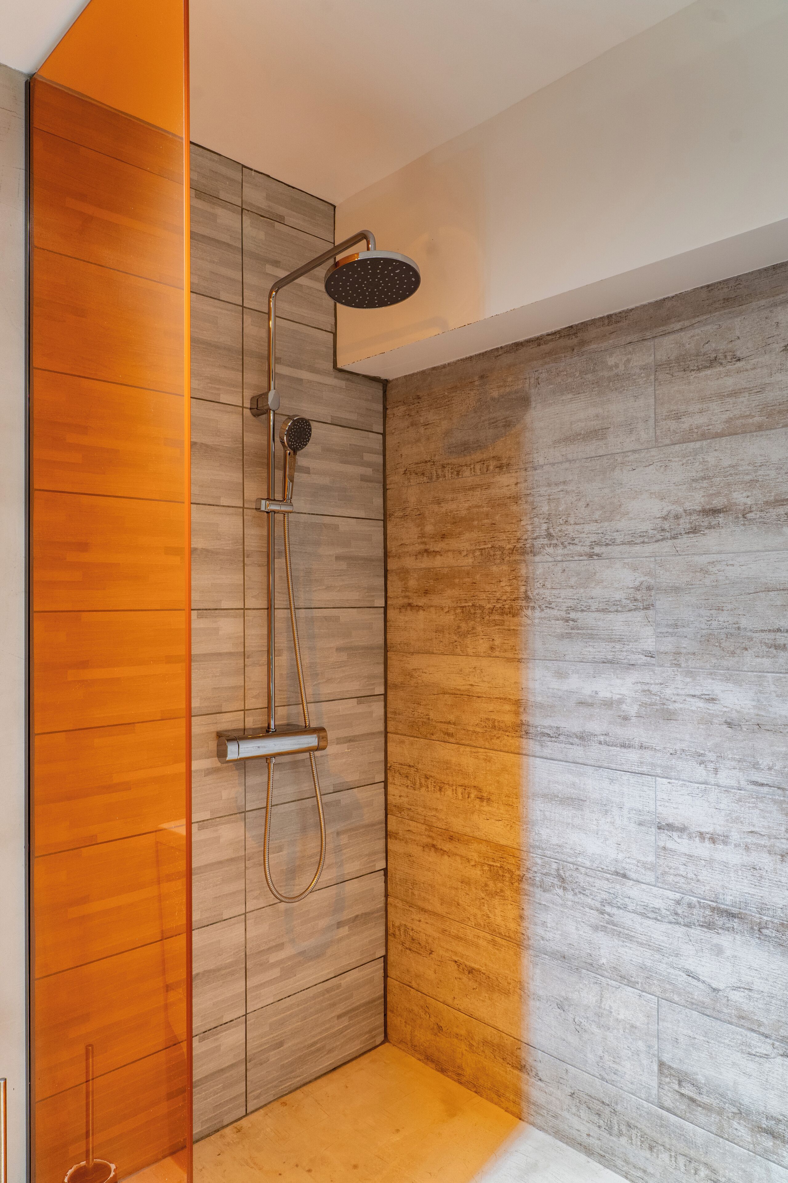 A modern shower with a rain showerhead and handheld showerhead, wooden paneling on the walls, and a tiled floor.