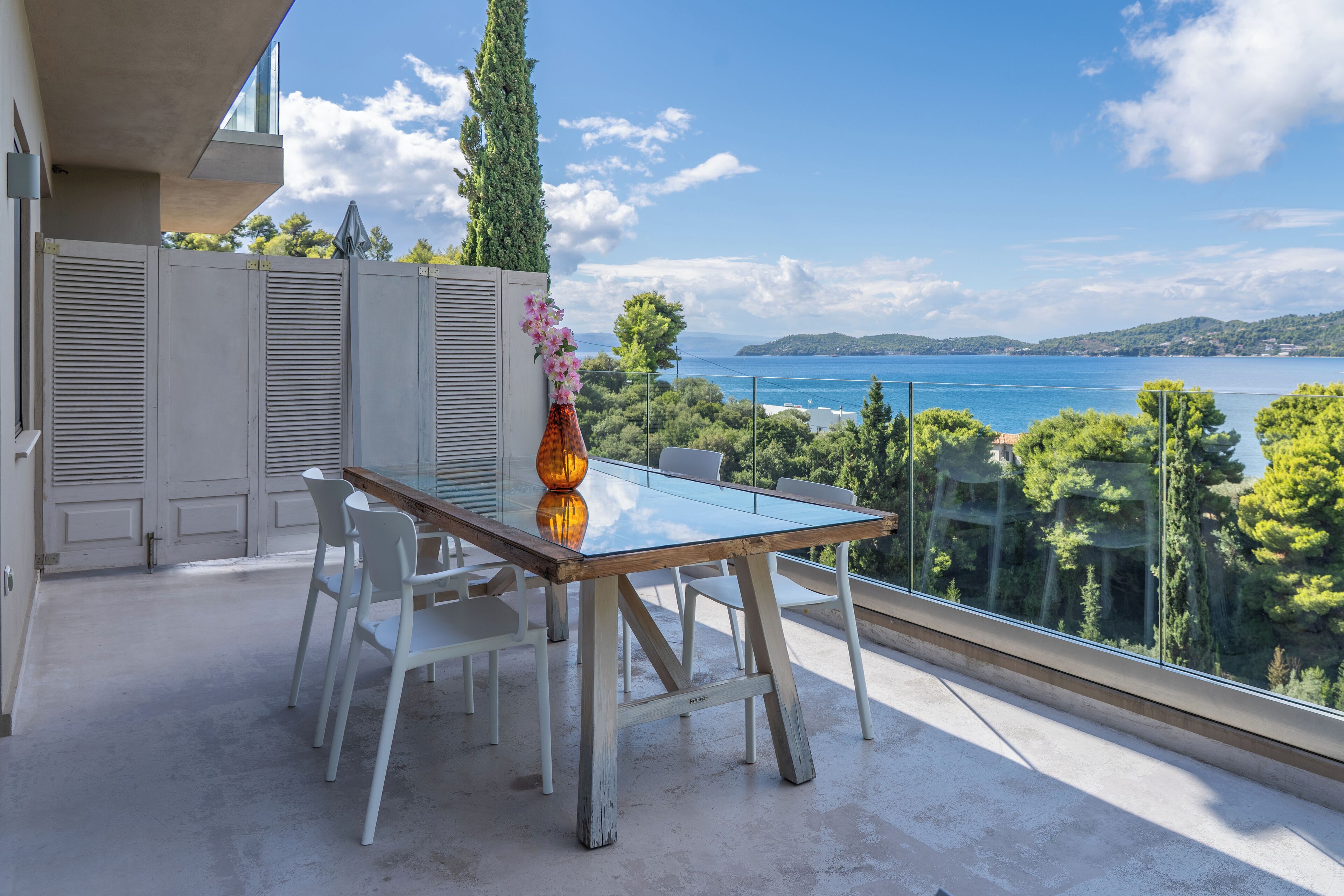 A balcony with a glass table and chairs, overlooking a scenic view of a body of water and greenery.