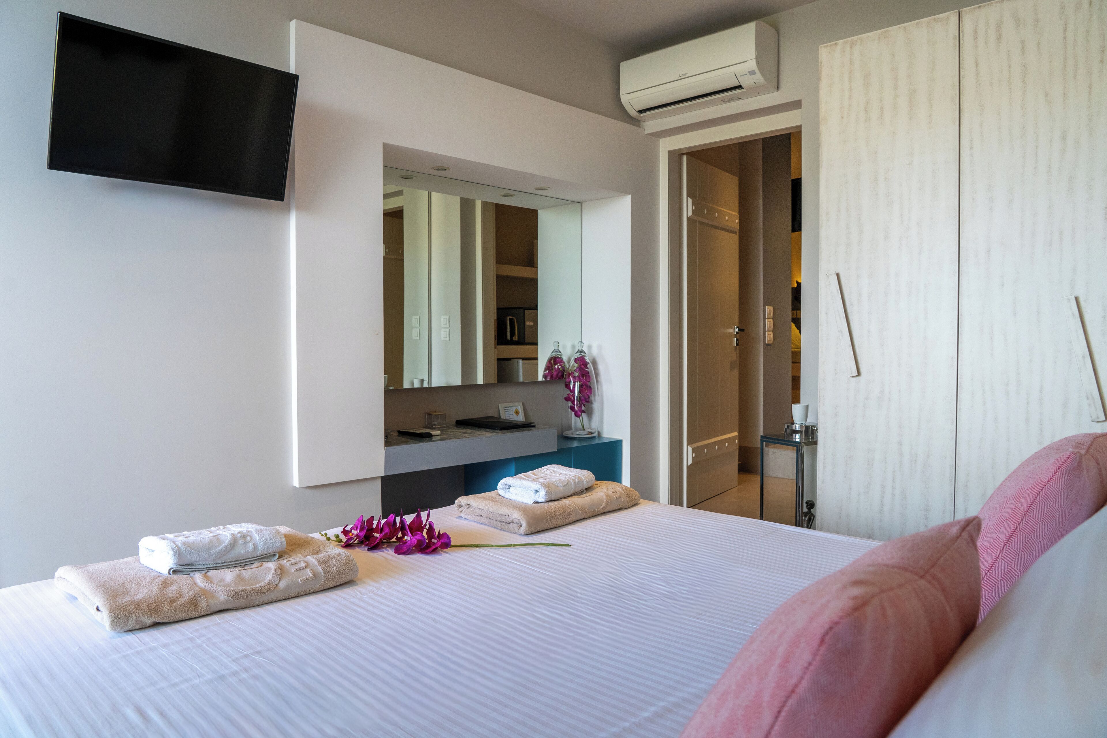 A hotel room with a bed, a flat-screen TV mounted on the wall, a mirror, a wardrobe, and an air conditioning unit.