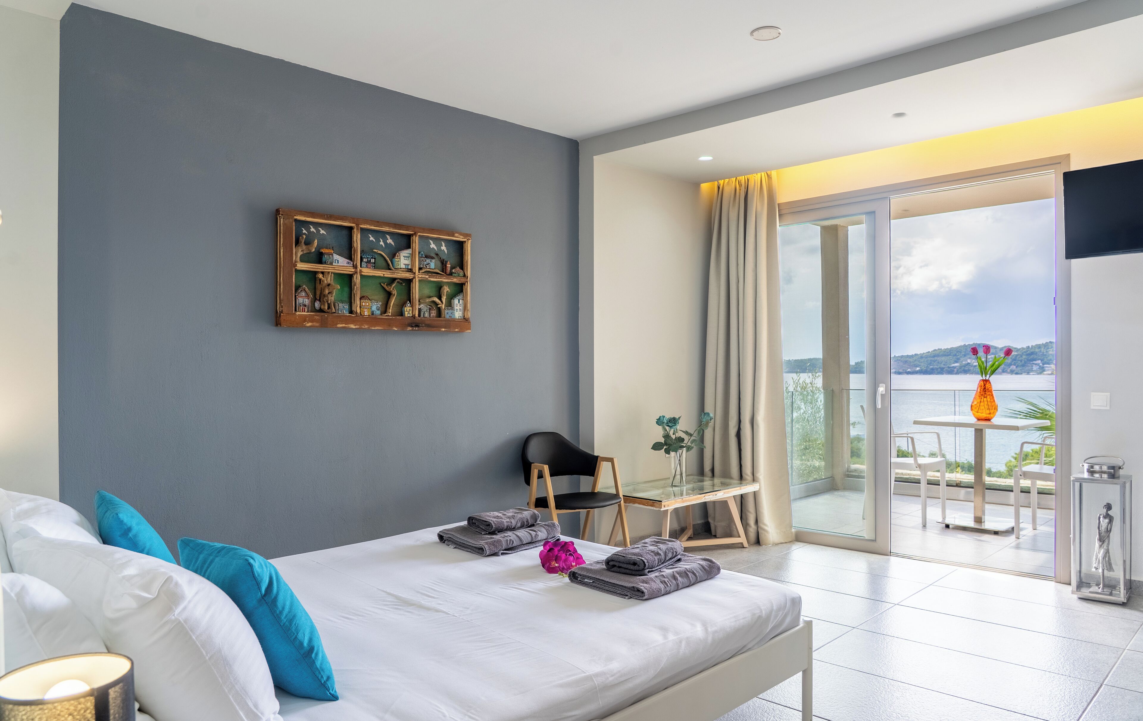 A modern bedroom with a bed, a small table with a chair, a balcony with a view, and a decorative wall piece.