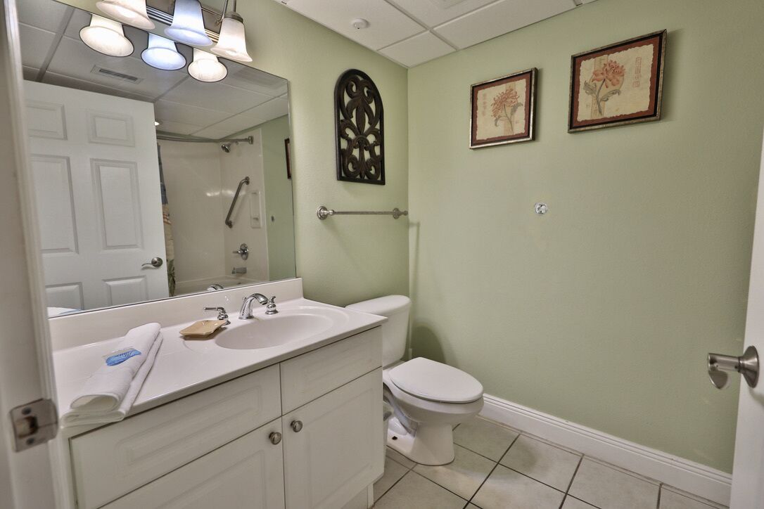 Ein Badezimmer mit einem weißen Waschbecken, Spiegel und Toilette. An der Wand hängen zwei gerahmte Bilder und über der Toilette ein dekoratives Metallobjekt.